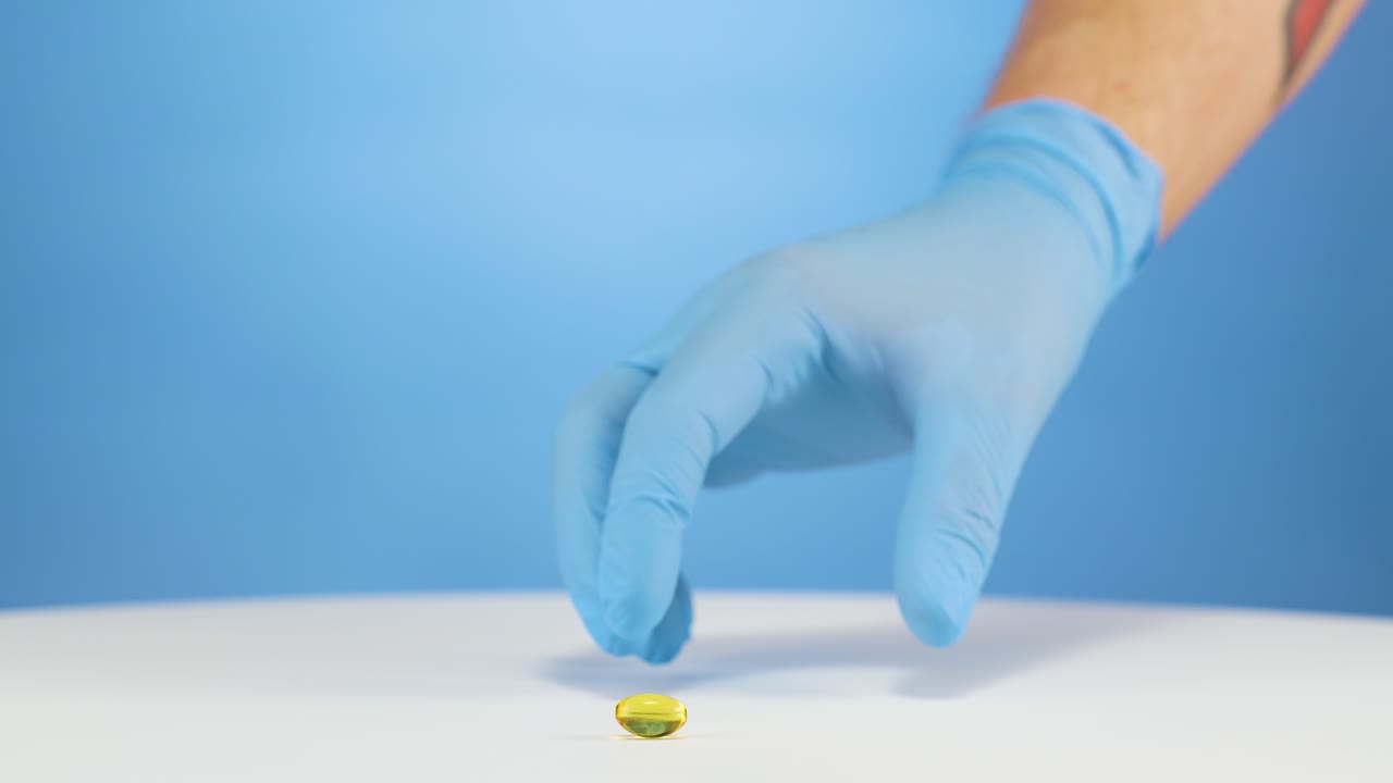 Mano caucasica con guanti blu che prende una singola capsula vegana di omega gialla, integratore
