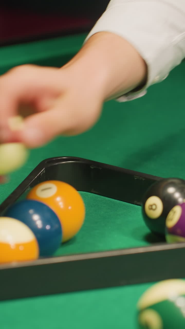 gentiluomo in camicia bianca che organizza le palle da biliardo usando un rack triangolare sul tavolo da biliardo verde. mani che allineano accuratamente le palle per una configurazione precisa. ripresa ravvicinata che cattura la preparazione focalizzata