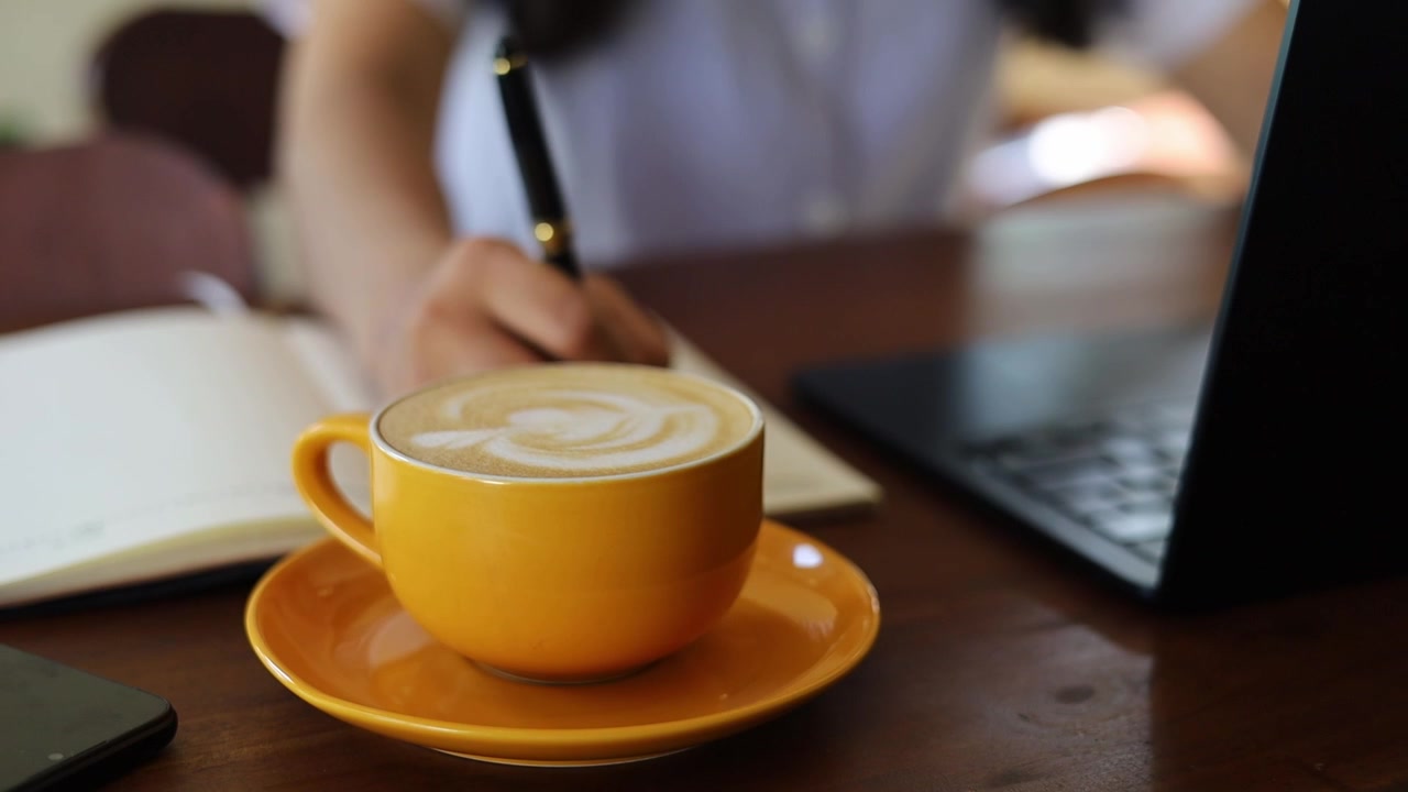 Mulher asiática está digitando no teclado do laptop, escrevendo em anotações, trabalhando em um café. Foco seletivo em uma xícara de café.