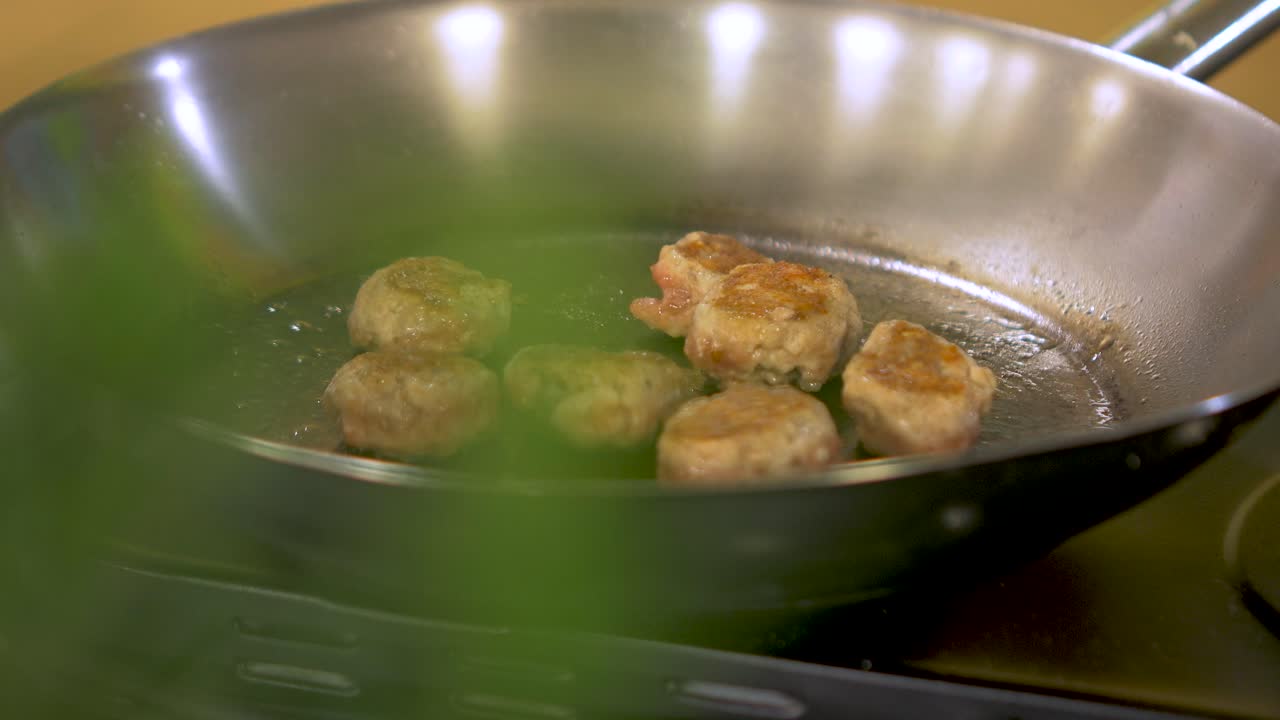 Small size juicy meat balls frying in a steel pan, handheld close up shot through out of focus green basil leaves