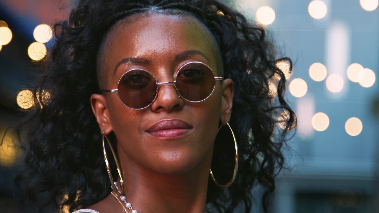 Young black woman looking up, turns to camera and takes off sunglasses, head shot, bokeh lights in background