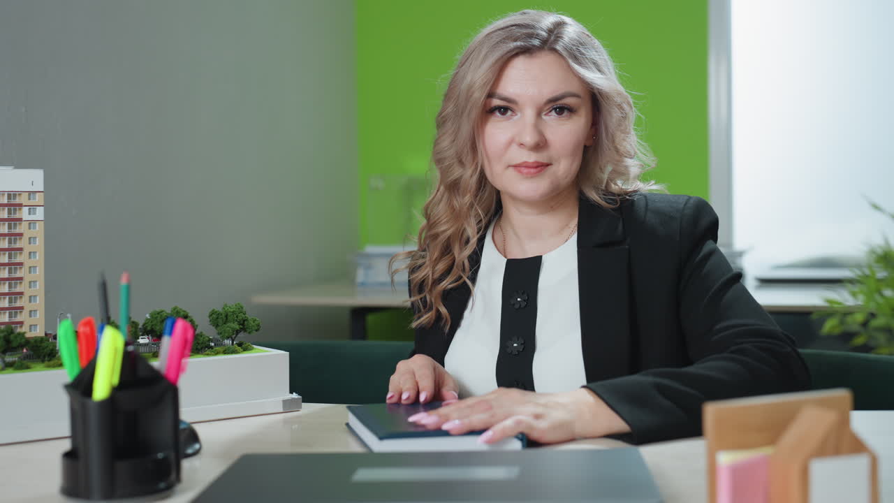 confident CEO closes book smiling posed at desk with architectural model laptop plant colorful pens in modern office setting professional charm and readiness organized layout