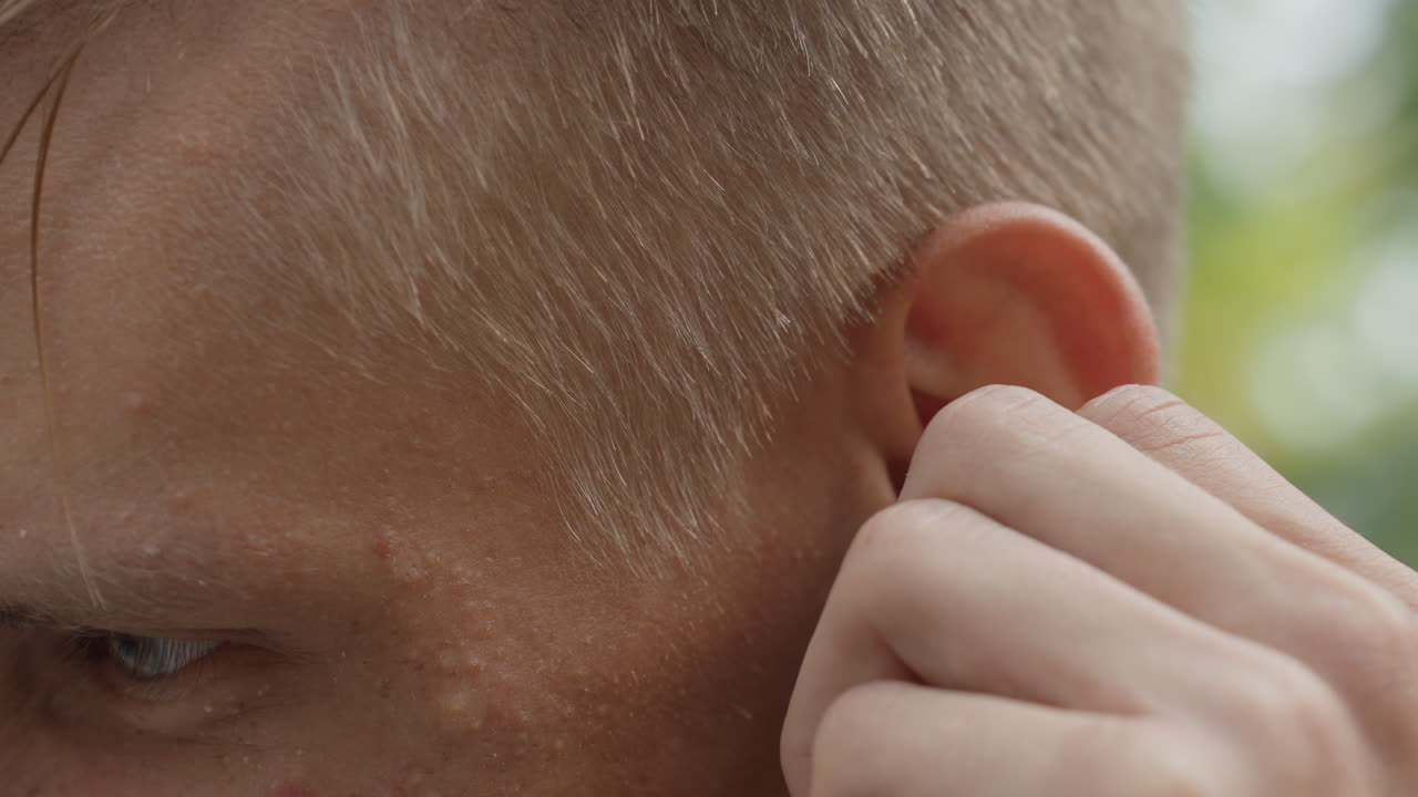 primo piano all'aperto di un uomo caucasico che si sistema un orecchino con la mano, profilo laterale che mostra pori e imperfezioni dell'acne, espressione concentrata, sfondo di un parco urbano, musicista o pendolare che si prepara, cura della persona spontanea