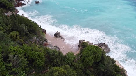 Aerial-view-of-Dhërmiu-Beach-in-Albania,-with-turquoise-waves-crashing-against-a-rocky-shoreline-surrounded-by-lush-greenery