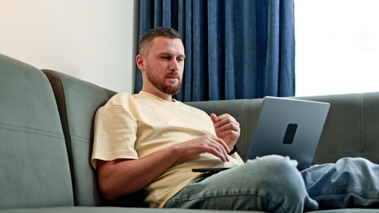 Man sits on couch using laptop in a room with blue curtains during daytime for work or study activity