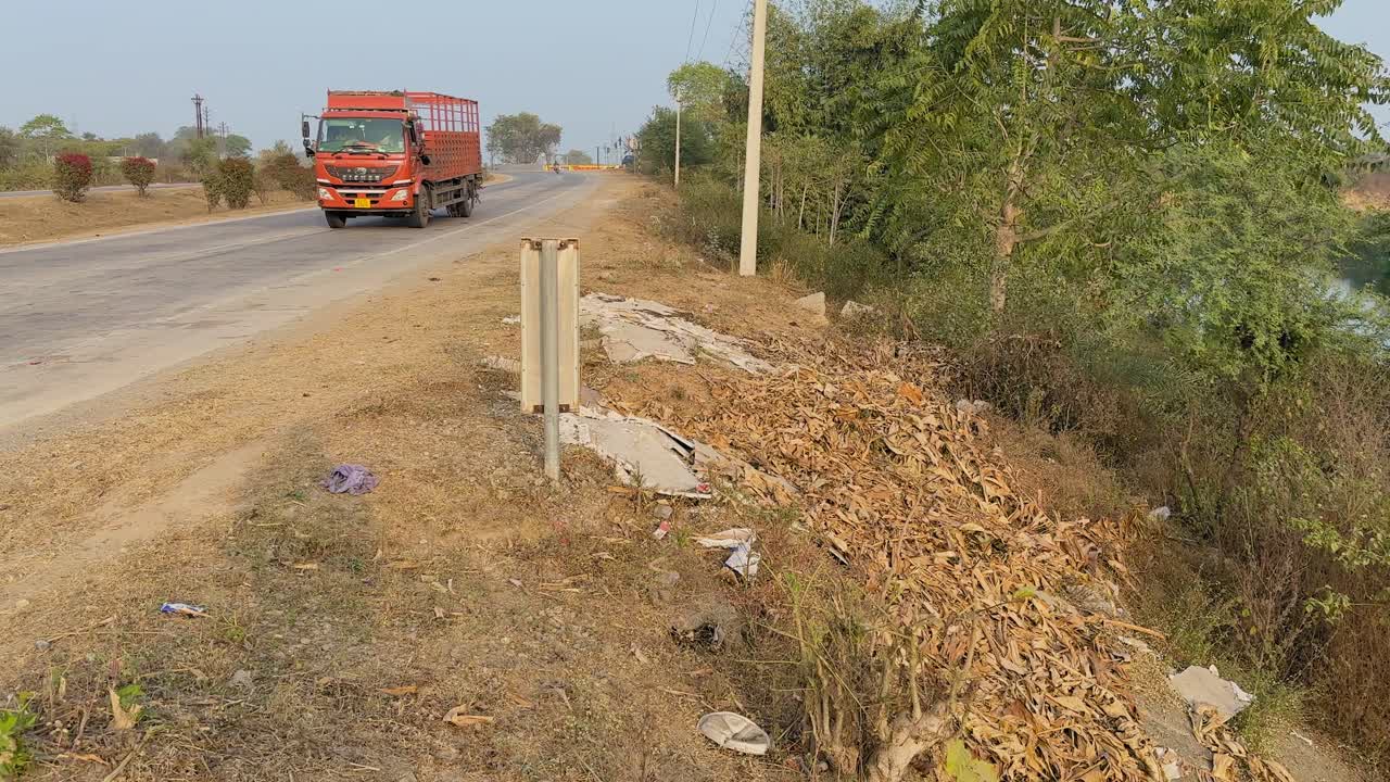 Spazzatura abbandonata sparsa lungo la banchina dell'autostrada accanto alla strada asfaltata, foglie secche e detriti ammassati vicino agli alberi mentre passano i camion, rivelando l'inquinamento stradale. Scena diurna