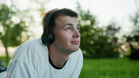 close up of man lying on grass wearing headphones nodding to music while using phone, gazing upwards with a thoughtful expression, surrounded by greenery and a soft bokeh effect in the background