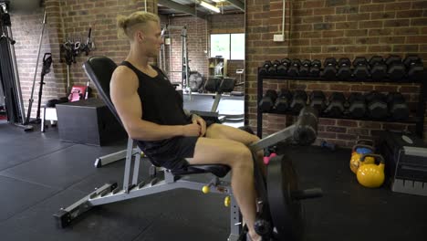 Hombre-Tatuado-En-El-Gimnasio-En-Casa-Sentado-En-Una-Silla-Haciendo-Extensión-Cuádruple