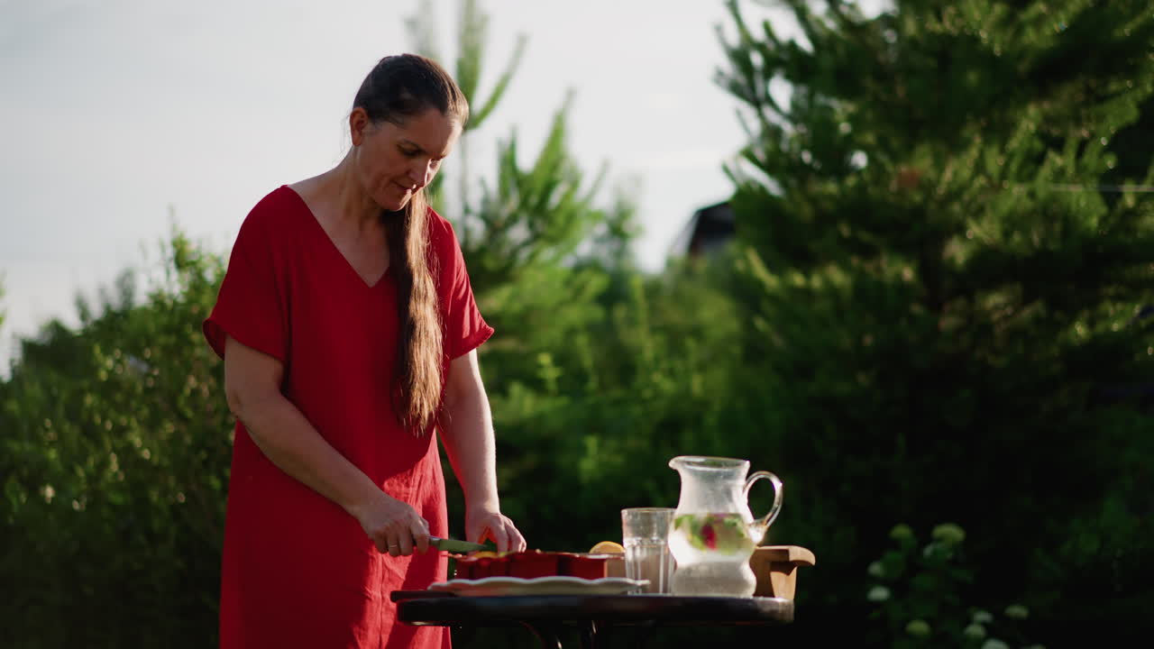 Donna in un giardino illuminato dal sole prepara una caraffa rinfrescante e frutta su un tavolo di legno, movimenti lenti e deliberati, calda luce dorata, sempreverdi con bokeh morbido, rituale domestico estivo, prodotti naturali e vetreria.