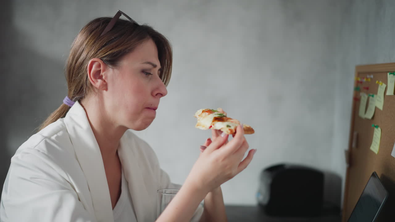 Freelancer with eyeglasses on head savoring slice of cheesy pizza while working remotely in cozy home office setting, sitting at desk with cork board full of sticky notes and laptop