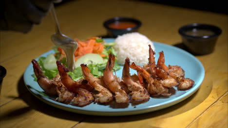 Cámara-Lenta-De-Una-Persona-Con-Un-Guante-De-Látex-Negro-Sirviendo-Una-Crema-De-Salsa-Blanca-Encima-De-Camarones-A-La-Parrilla-Con-Ensalada-Y-Arroz-Blanco