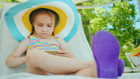 Una-Niña-Linda-Con-Un-Gran-Sombrero-Multicolor-En-Traje-De-Baño-Está-Sentada-En-Una-Tumbona-Y-Jugando