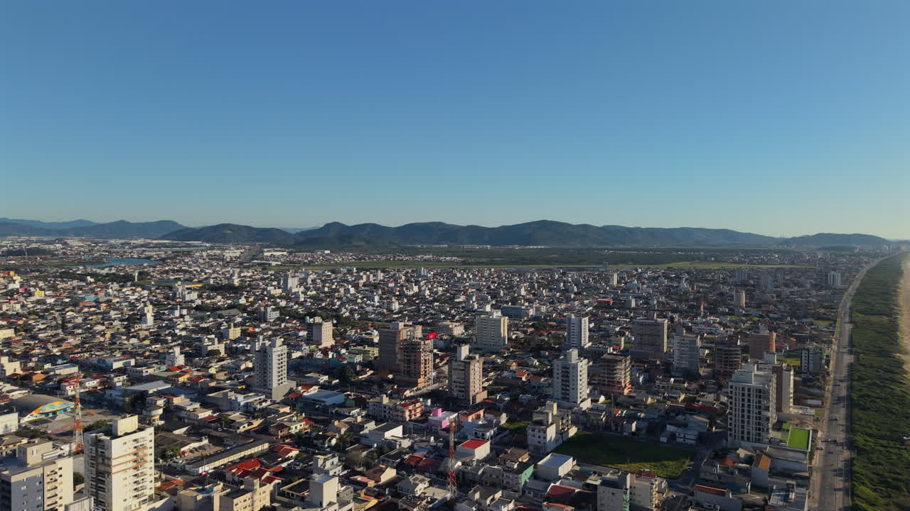 Drone flies upward and forward over Navegantes, Brazil, showing residential areas, high-rises, beach, dunes, a coastal road, sea, and distant mountains