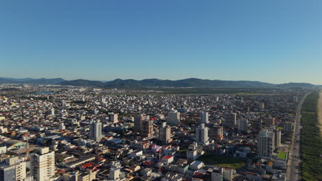 drone flies upward and forward over navegantes, brazil, showing residential areas, high-rises, beach, dunes, a coastal road, sea, and distant mountains