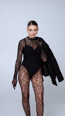 rear view of a woman wearing black bodysuit and lace long sleeve and stockings holds a jacket. model poses walking in studio on high heels. vertical video.