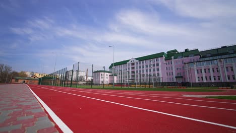 approaching a soccer field surrounded by fence in front of the big pink building. red running track at the foreground. low angle view.