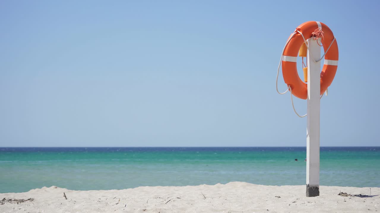 오렌지색 구명보이가 파란 하늘과 푸른 바다를 배경으로 모래 해변의 기둥에서 매달려 있습니다.