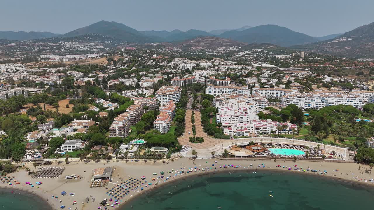 A sweeping aerial view of Marbella’s coastline reveals sandy beaches, turquoise waters, and a lively townscape backed by rolling mountains under a clear sky