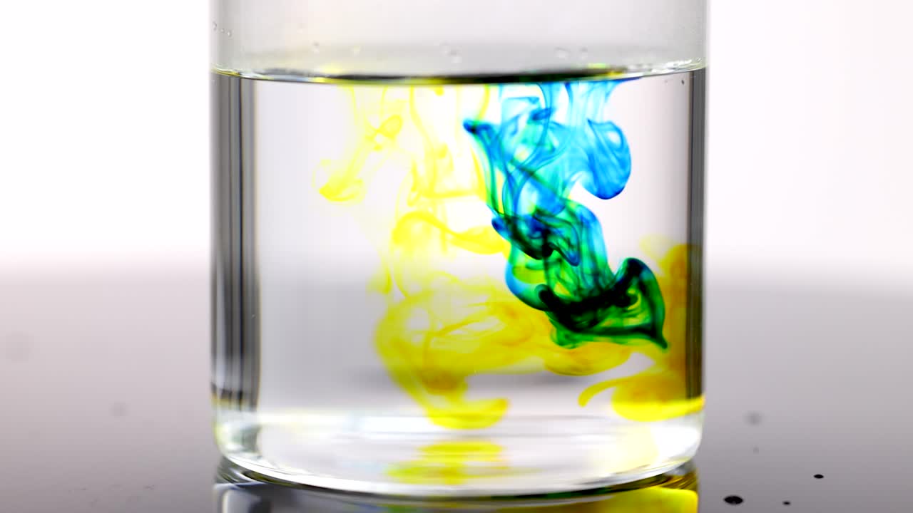 Blue and yellow liquid dyes swirl and blend in water under bright studio lighting