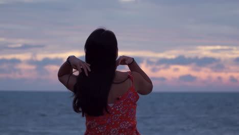 Asian-woman-standing-by-the-ocean-in-tropics-admiring-sunset-with-dramatic-sky-and-touching-her-long-hair,-back-view