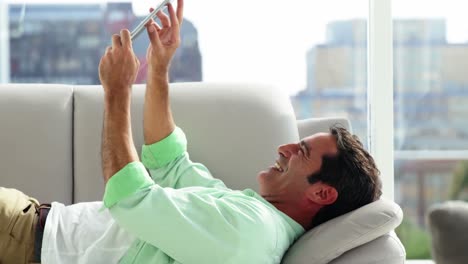 Smiling-man-lying-on-sofa-and-using-digital-tablet-in-living-room-at-home