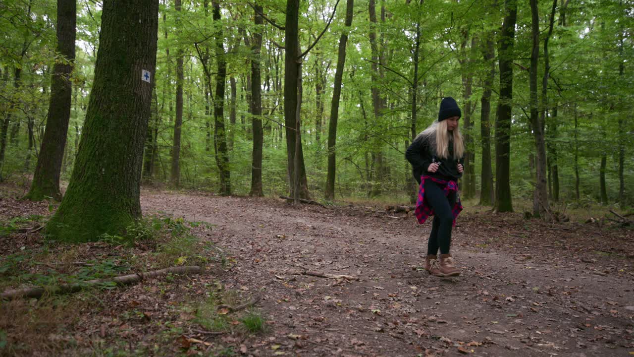 A woman with a backpack walks along a dirt path surrounded by tall green trees in a calm forest. The scene captures the peaceful atmosphere of an autumn hike and connection with nature