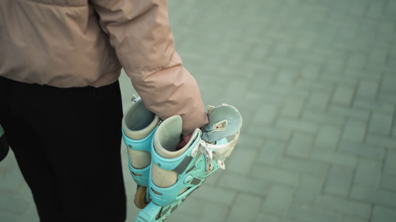 Close-up shot of a girl dressed in a peach jacket and black trousers, carrying a pair of rollerblades as she walks along a paved park path during sunset.with the girl's face not visible