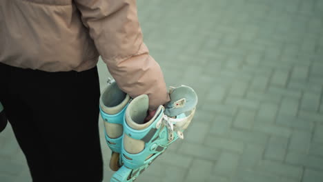 Close-up shot of a girl dressed in a peach jacket and black trousers, carrying a pair of rollerblades as she walks along a paved park path during sunset.with the girl's face not visible