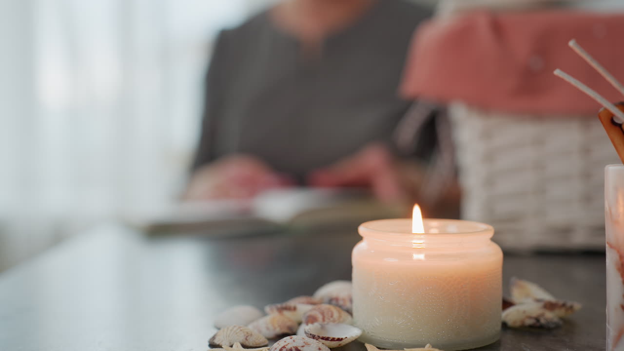 Close up of candlelight burning on table surrounded by seashells with blurred view of basket and person flipping through book in cozy softly lit indoor setting filled with warmth calmness