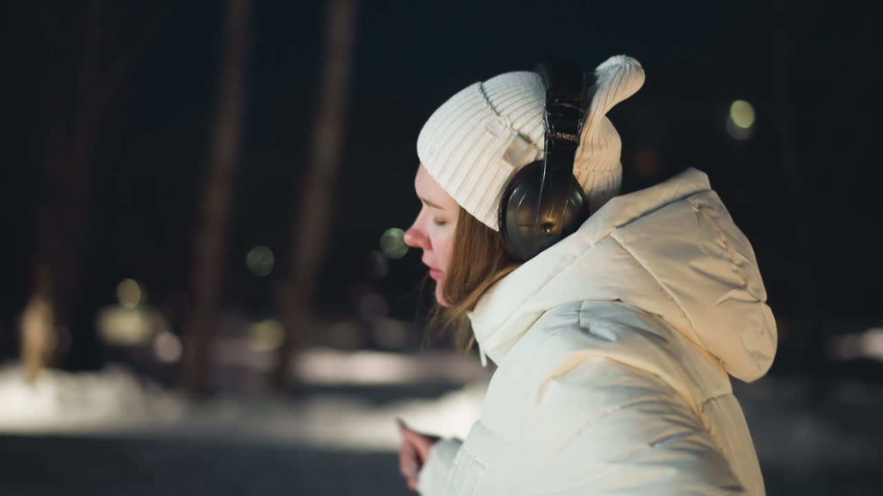 Side view of snow dancer moving her body passionately and hands in cool evening breeze wearing headphones with smile on face under soft ambient park lights on snow covered walkway at night