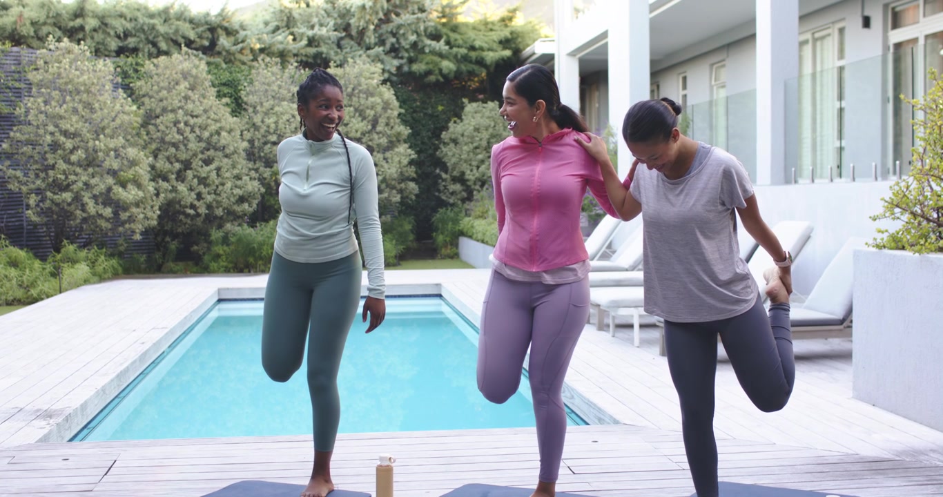 Donne diverse in leggings che si tengono le caviglie su tappetini da yoga a bordo piscina, quella al centro si stabilizza a destra per l'equilibrio.