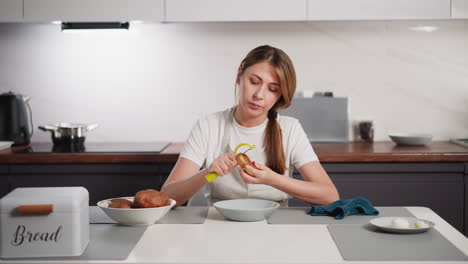 housewife wearing apron seated at kitchen table with head slightly slanted while using peeler to remove skin from raw potato, surrounded by bowl of potatoes, boiled eggs, and kitchen items