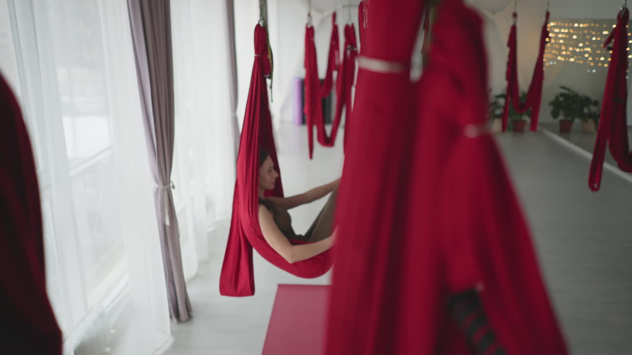 Bright yoga studio with tall windows and sheer curtains as woman lies comfortably in red silk hammock swinging gently with eyes closed smiling conveying serene wellness and balanced flow