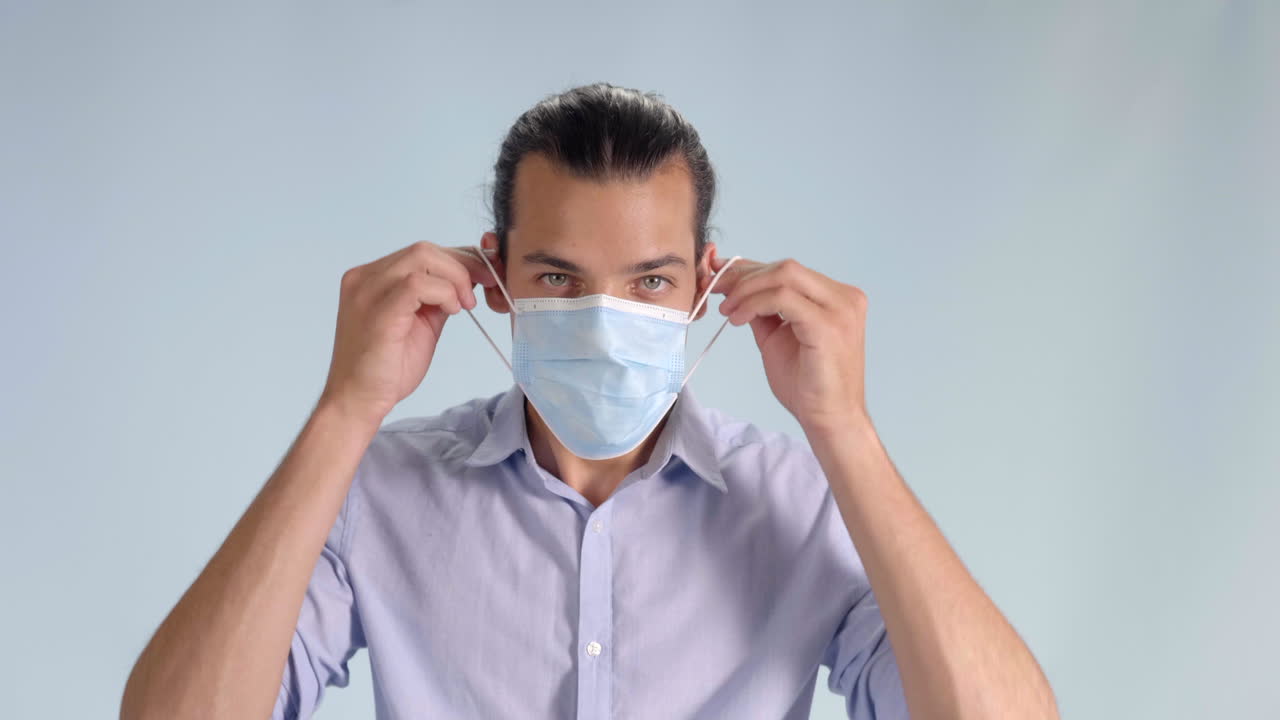 깃이 달린 버튼 다운 셔츠를 입은 개암색 눈을 가진 행복한 성인 남성은 covid-19 - 코로나바이러스로부터 보호하기 위해 회색 배경에 외과용 마스크를 착용하고 삼각대를 가까이서 촬영합니다.