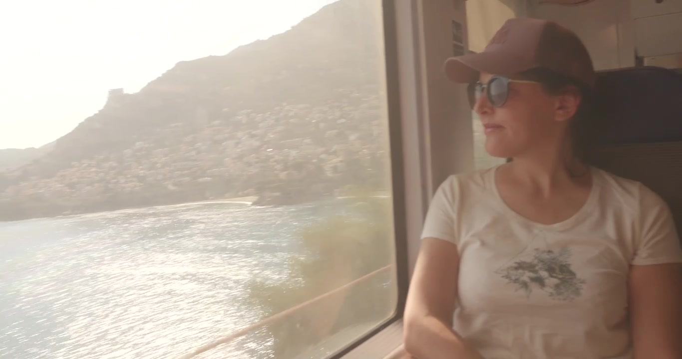 Passeggera con berretto e occhiali da sole siede accanto a un finestrino del treno osservando la costa della Costa Azzurra che scorre sotto la luce estiva