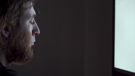 man with beard in dimly lit office late night working computer screen rubs face tired and alone