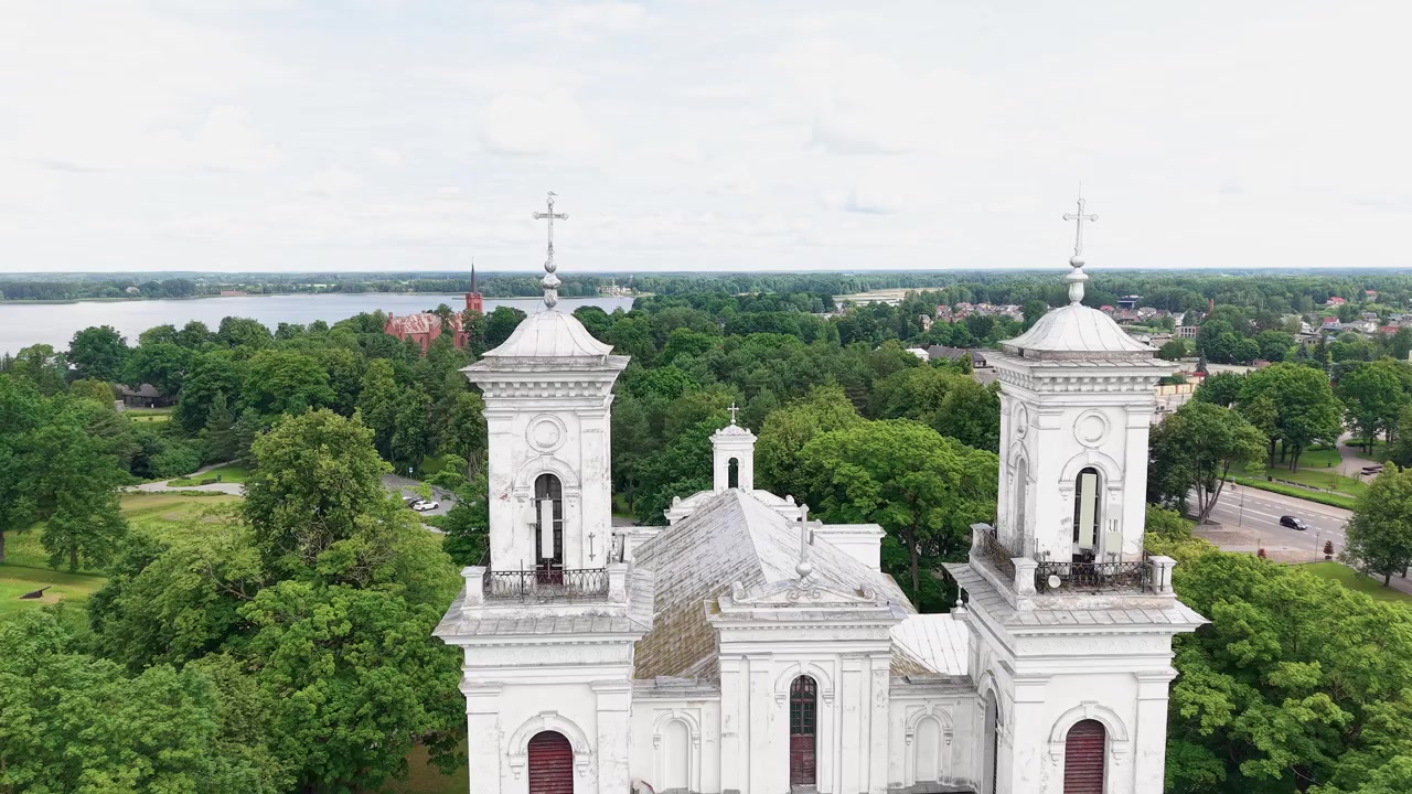 Splendide riprese da drone catturano le torri bianche della Chiesa di San Giovanni Battista di Biržai tra alberi verdi, sullo sfondo di una suggestiva città e lago lituani sotto una luce diurna brillante e diffusa.