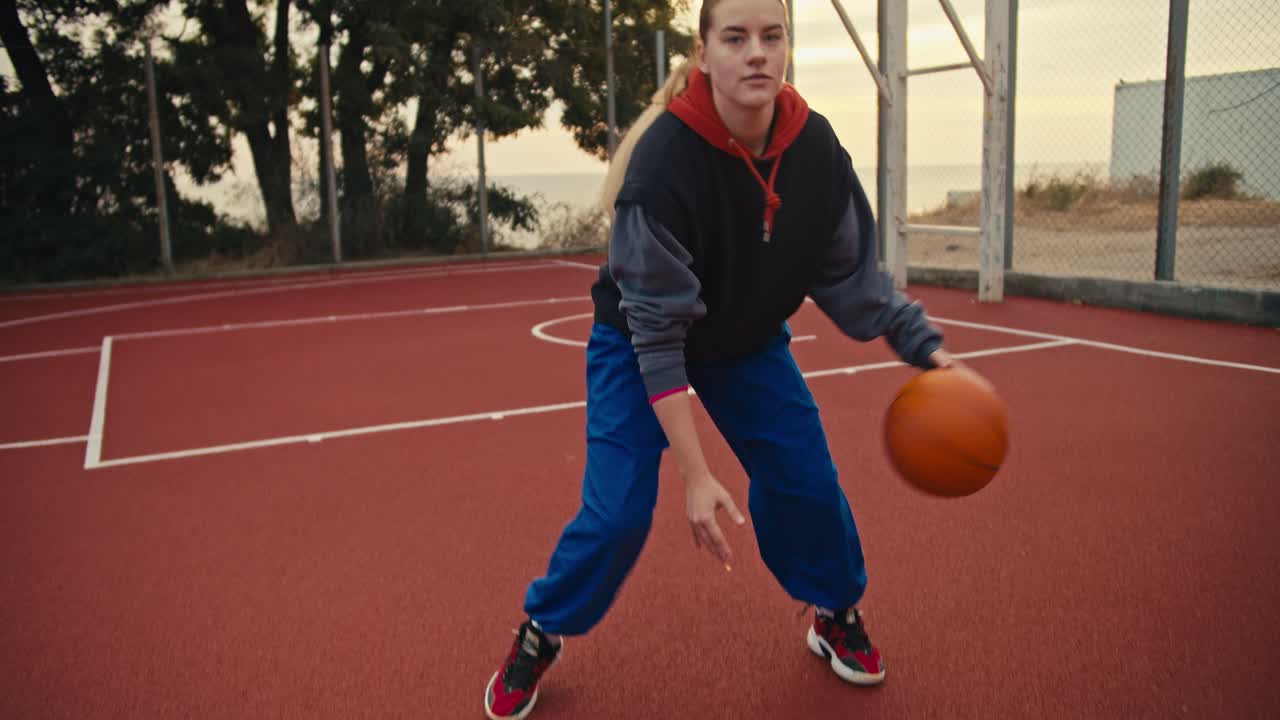 retrato de cerca una chica rubia en pantalones azules golpea la pelota y luego posa y mira a la cámara durante su práctica matutina en la cancha de baloncesto roja por la mañana