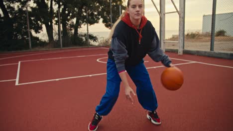 Close-up-Portrait-a-blonde-girl-in-blue-pants-hits-the-ball-and-then-poses-and-looks-at-the-camera-during-her-morning-practice-on-the-red-basketball-court-in-the-morning