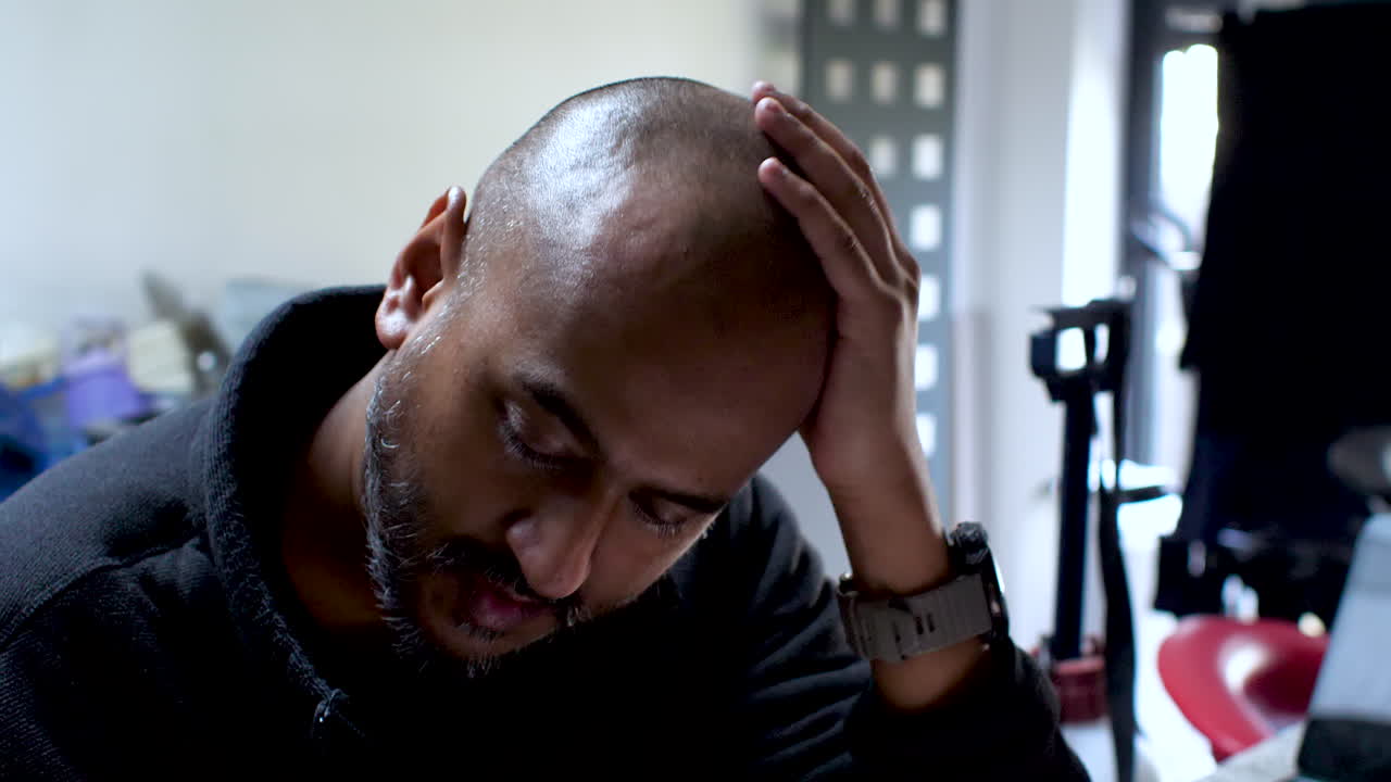 Bald South Asian British Male sits rubbing hands over head, his expression fraught with the weight of burdens, vividly capturing the palpable strain of stress and anxiety