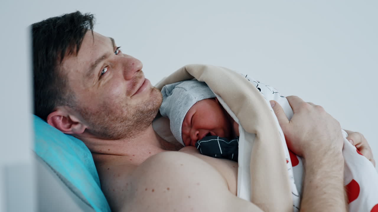 Dark-haired Caucasian man lies on the couch at hospital. Father is holding his crying newborn child on the chest for skin to skin contact.
