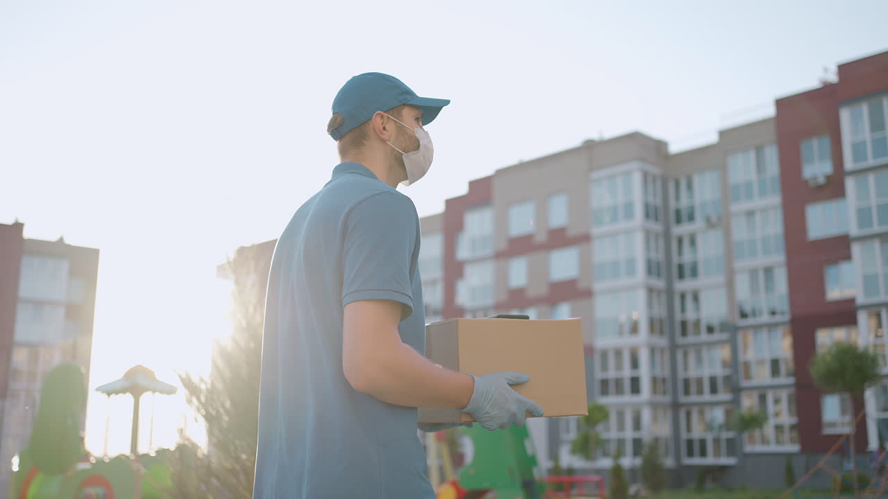 el repartidor masculino con una gorra y una máscara protectora y guantes va con una caja en sus manos y lleva un paquete al cliente. entregando pedidos a domicilio en línea.