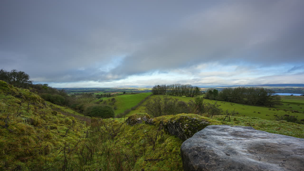 timelapse del paisaje de tierras de cultivo de naturaleza rural con rocas y hierba en primer plano y árboles en la distancia durante el día nublado soleado visto desde carrowkeel en el condado de sligo en irlanda