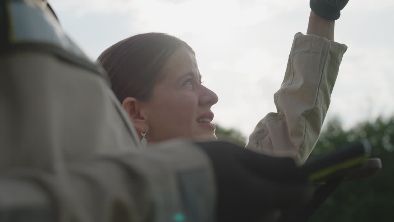 close up of gloved hand shading eyes of woman holding mobile device while colleague shoulder enters frame, sunlight diffused through clouds above grassy background