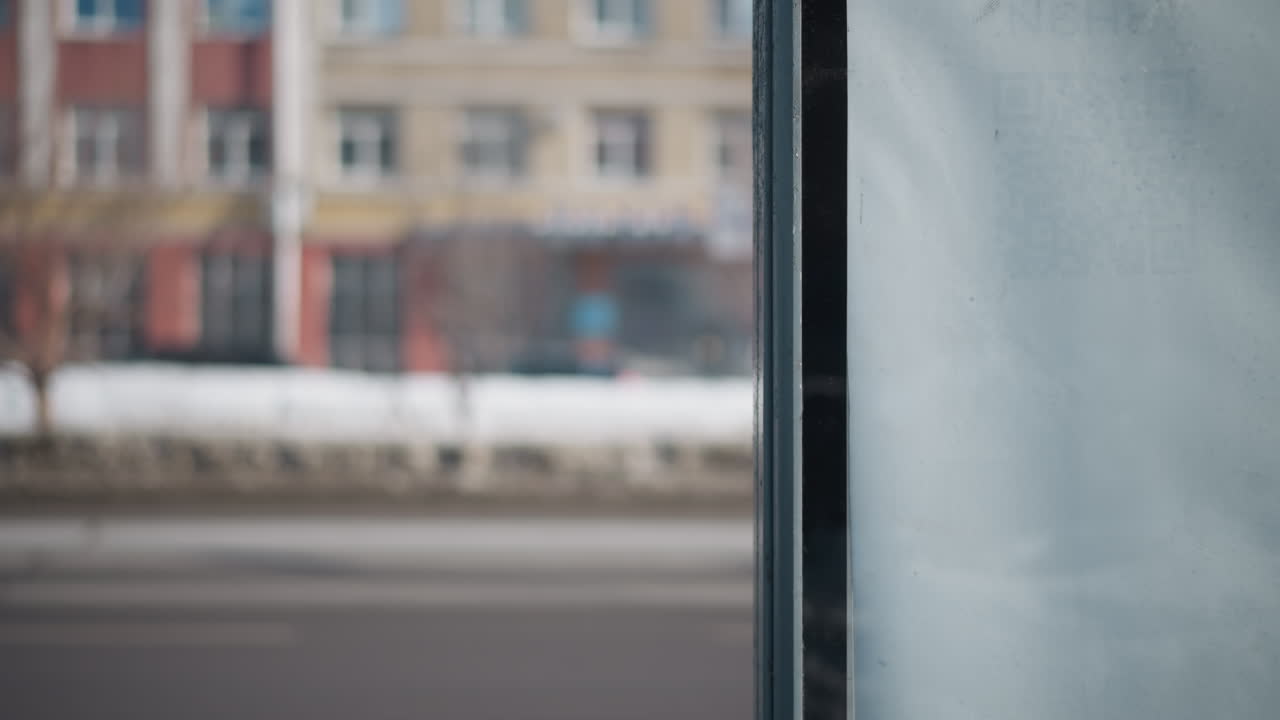 transparent window view shows person in winter coat passing along city pavement while cars move on snowy road, blurred buildings beyond, cold commute mood with frosted pane and soft motion