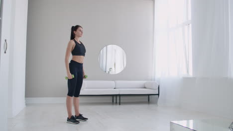 A-woman-raises-her-arms-with-dumbbells-doing-a-shoulder-exercise-in-her-light-white-apartment-against-a-sofa-and-window.