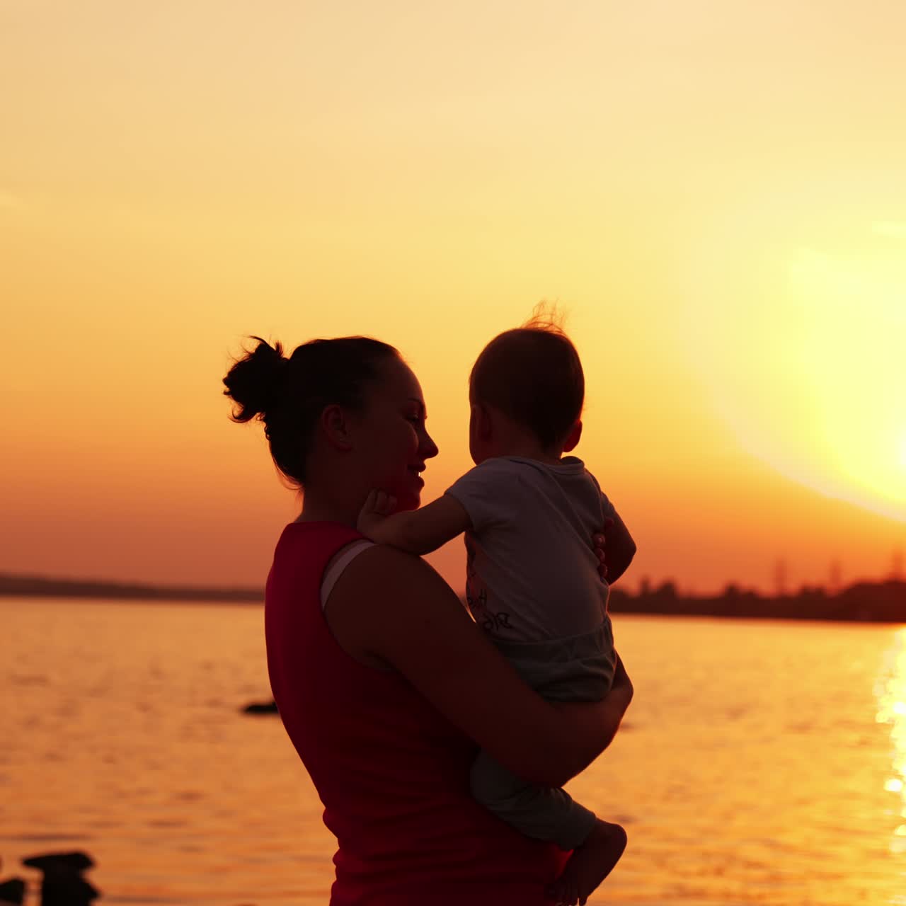 Brunette woman in red sleeveless shirt holding a little toddler. Mother and son enjoying beautiful sunset near the river
