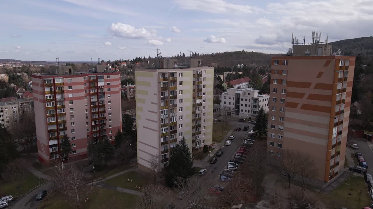 An aerial view of three residential apartment buildings surrounded by greenery and a nearby forest. The neighborhood consists of various houses, roads, and parked cars, creating a peaceful atmosphere