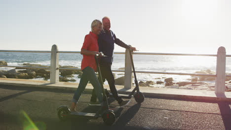 Senior-couple-walking-next-to-electronic-scooter-alongside-beach