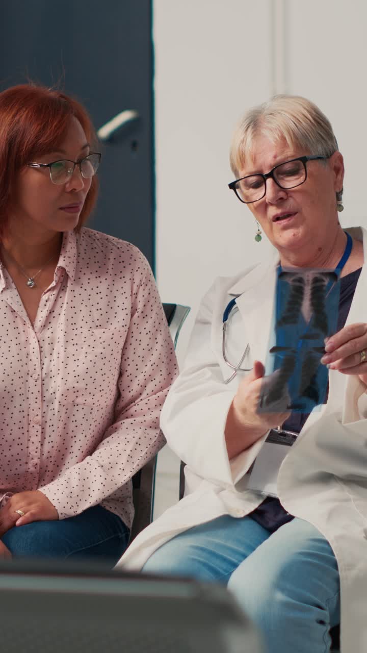 Vertical video: Senior medic and asian woman analyzing x ray scan in clinic lobby